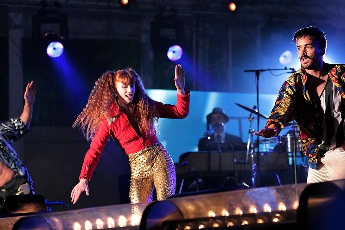 Livemuziek. Met een drummer, gitarist en dansende vrouw. Ze dragen felgekleurde kleding en staan op een podium waar meerder spotlights op gericht zijn.