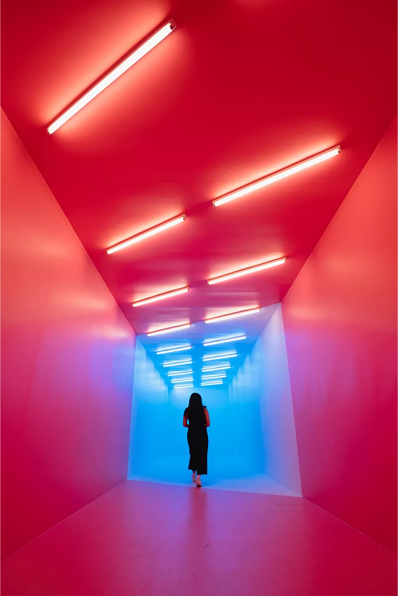 Persoon loopt door een gang. Aan het plafond hangen tl-buizen. De gang en het licht gaat van rood naar blauw,